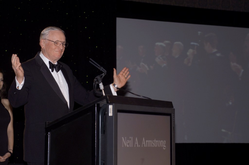 Neil Armstrong, the first person to walk on the Moon, stands at a podium during a formal event. He is wearing a black tuxedo with a bow tie, and he is smiling with his arms slightly raised.