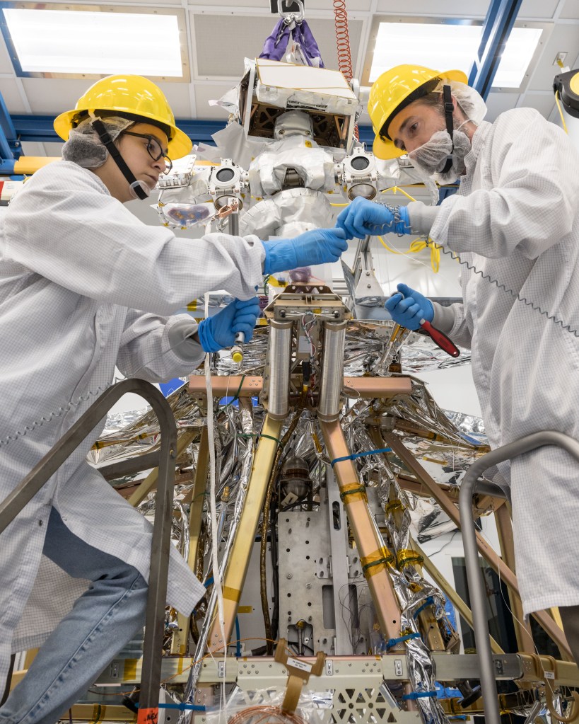 Two engineers work on a complex piece of equipment in a cleanroom environment. Both are wearing white lab coats, yellow hard hats, blue gloves, and face masks or beard covers.