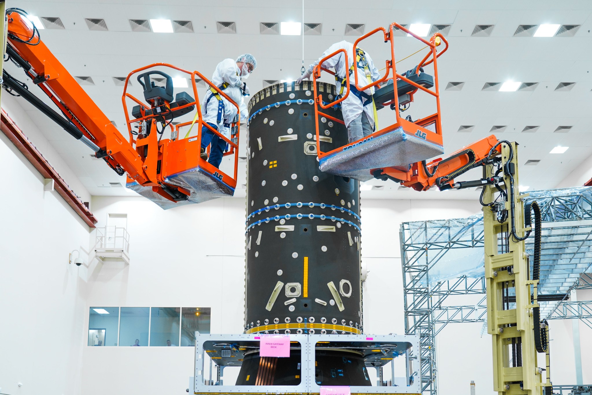 Two engineers in cleanroom suits work from elevated orange platforms on the cylindrical structure of the Power and Propulsion Element (PPE) at Maxar Space Systems in Palo Alto, California.