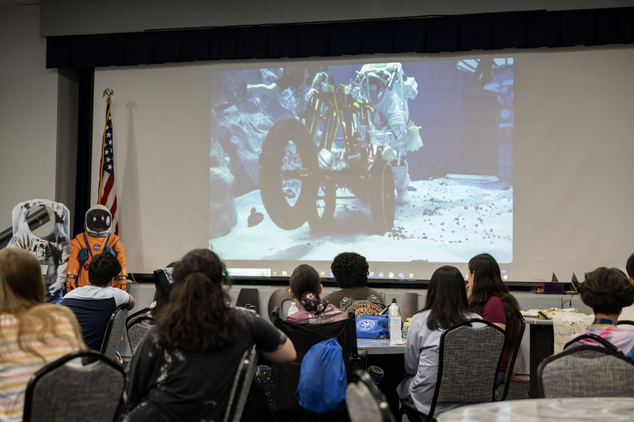 High school students sit with their backs to the camera as they watch a large screen displaying a white extravehicular activity suit being tested via virtual classroom