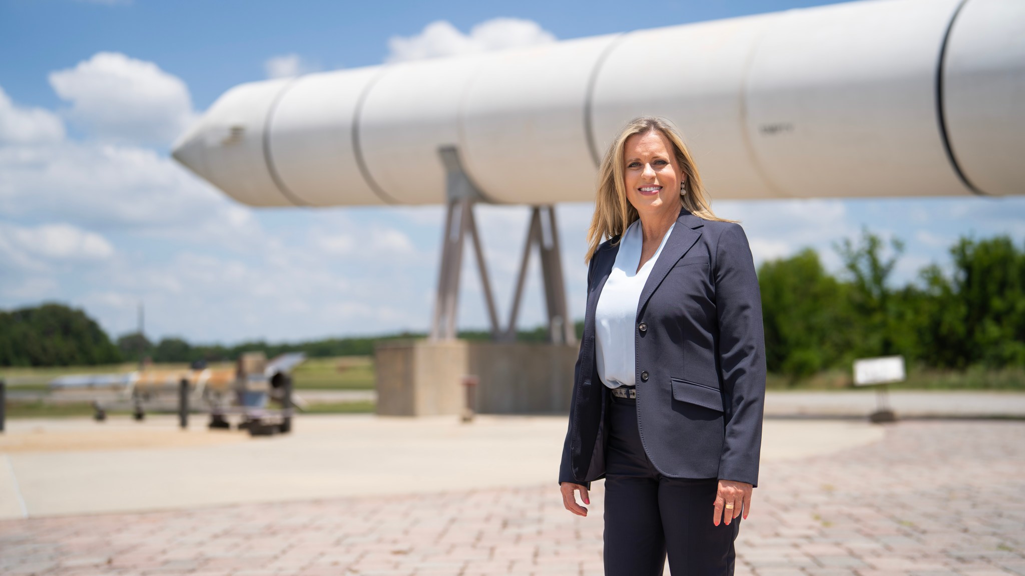 A blonde woman stands in front of a rocket booster in a grey jacket.