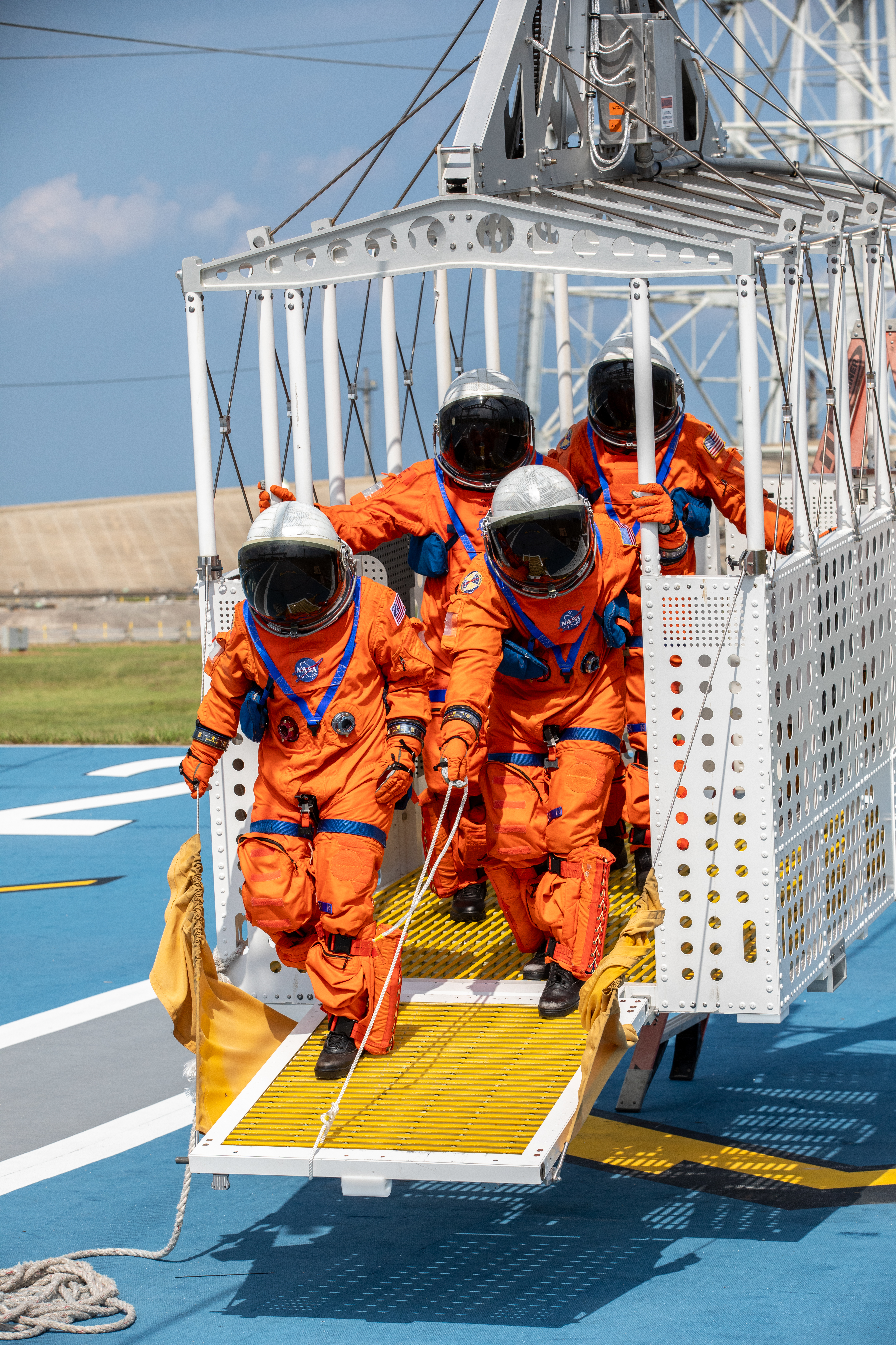 Four people dressed in orange astronaut suits and white helmets with black visors begin to get out of a large white metal basket that is lifted some inches above the ground.