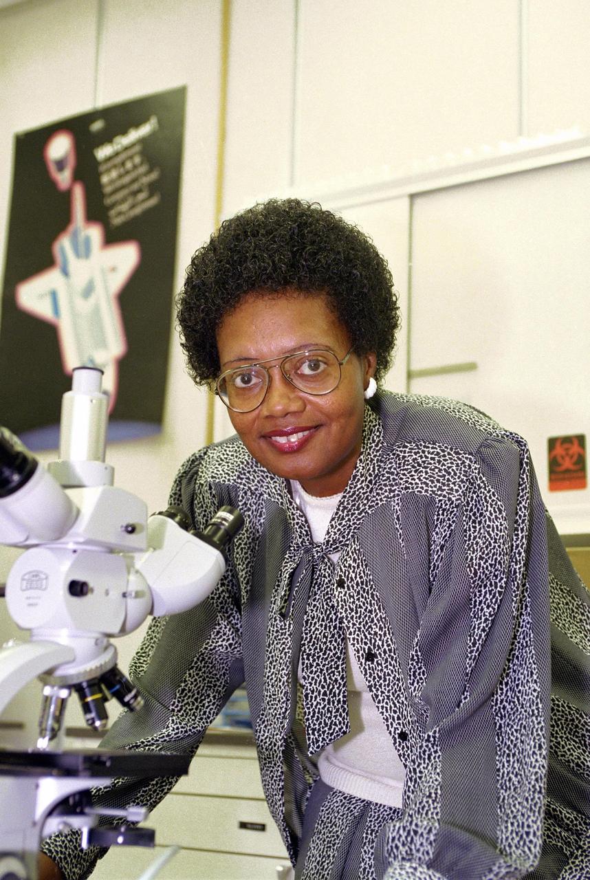 Dr. Long, an African American woman with curly hair, poses for a picture while leaning over a white microscope. She wears large aviator-shaped glasses and a black and white patterned top and pants.