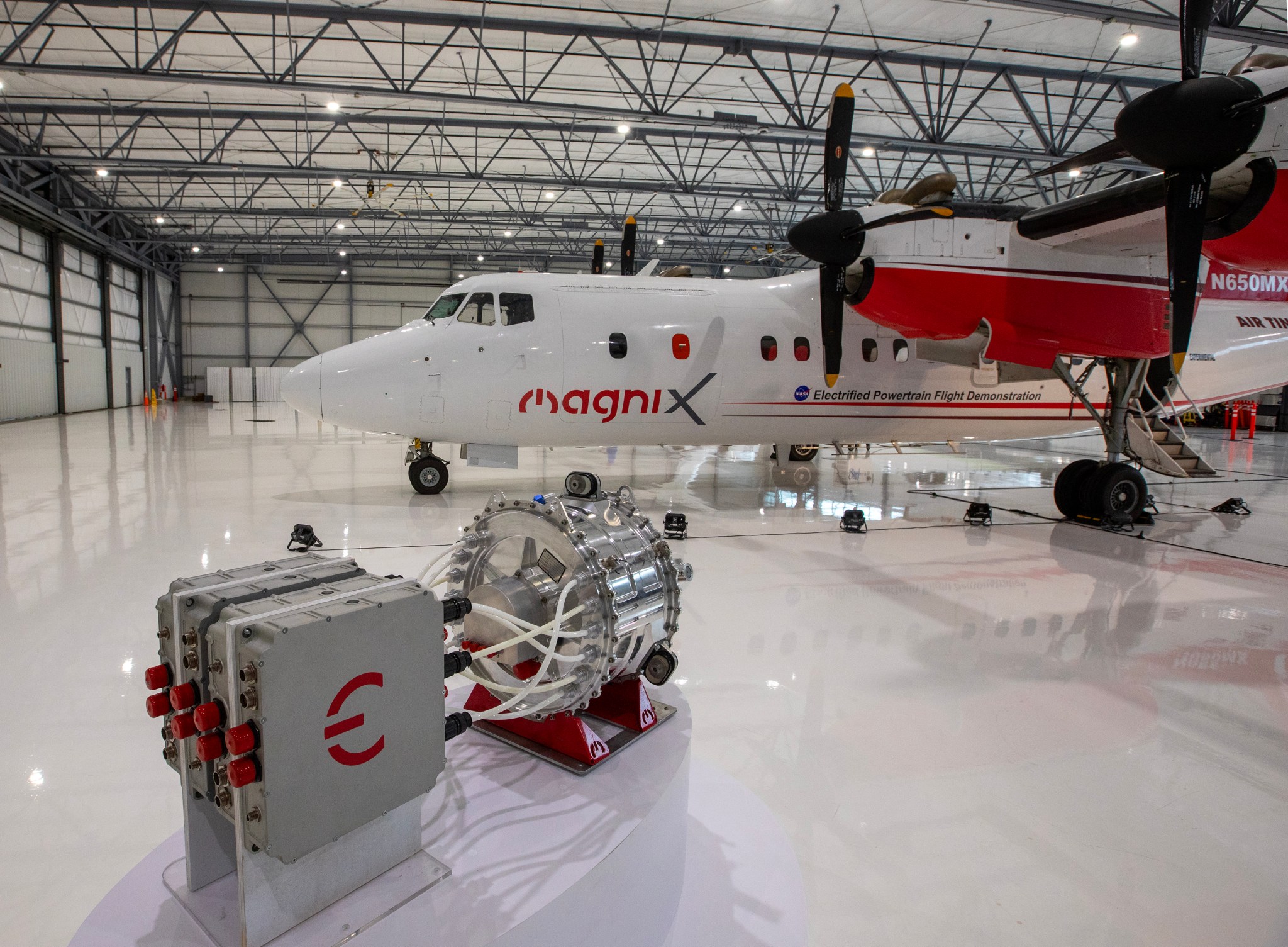 A four engine turboprop aircraft wrapped in a red and white livery with logos and names of each partner on the project sits under the lights inside an aircraft hangar. On the ground in front of the plane is an electric powertrain with an electric motor and battery pack that will soon be swapped out with one of the aircraft’s traditional engines to form a hybrid electric system.