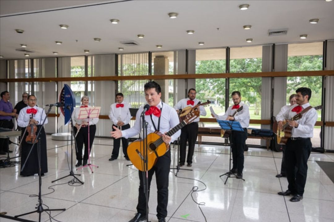 A group of musicians, dressed in traditional mariachi attire with white shirts, red bow ties, and black pants, perform in a bright, spacious room. The lead musician stands at the front, holding a guitar and singing into a microphone.