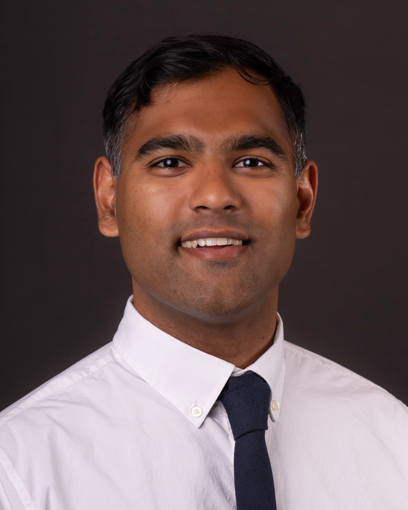 Headshot against a black background. Tyler Parsotan wears a wears a white shirt with a black tie.