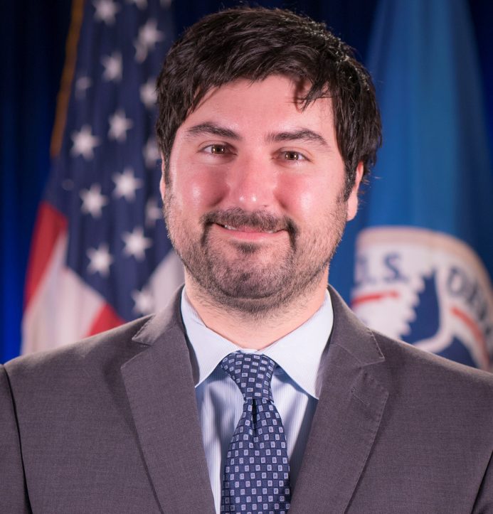 Official headshot of Matthew Dosberg, smiling in a suit in front of flags.