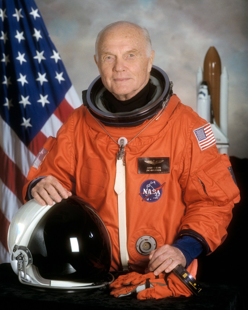 A formal portrait of astronaut John Glenn, dressed in an orange NASA space shuttle flight suit. He is holding a white and black helmet in one hand and has his other hand resting on a pair of orange gloves.