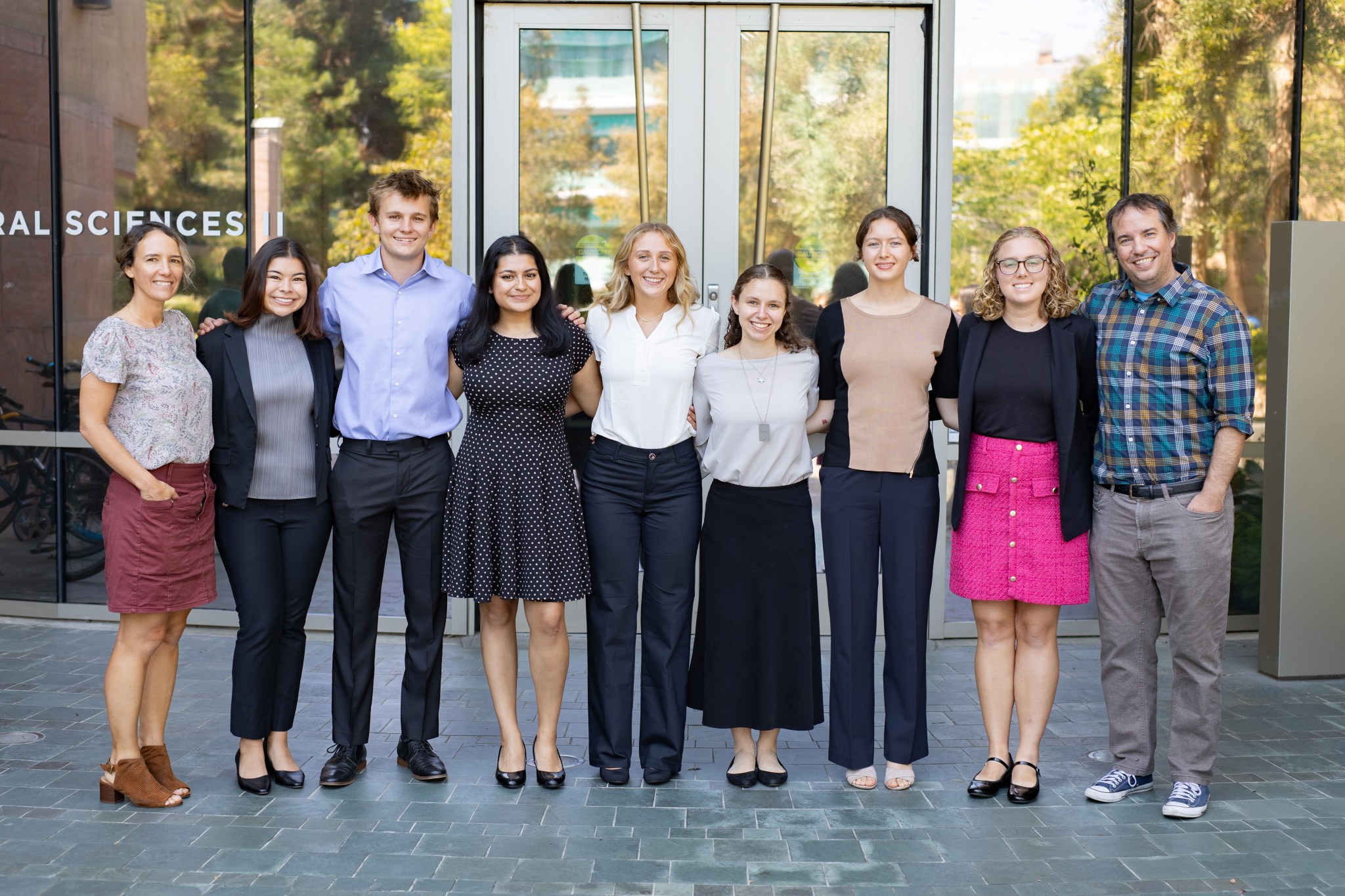 A group of nine people. the first eight college age and the last one a professor, stand in a line in professional attire. Behind them is a glass building with glass doors, reflecting green trees.