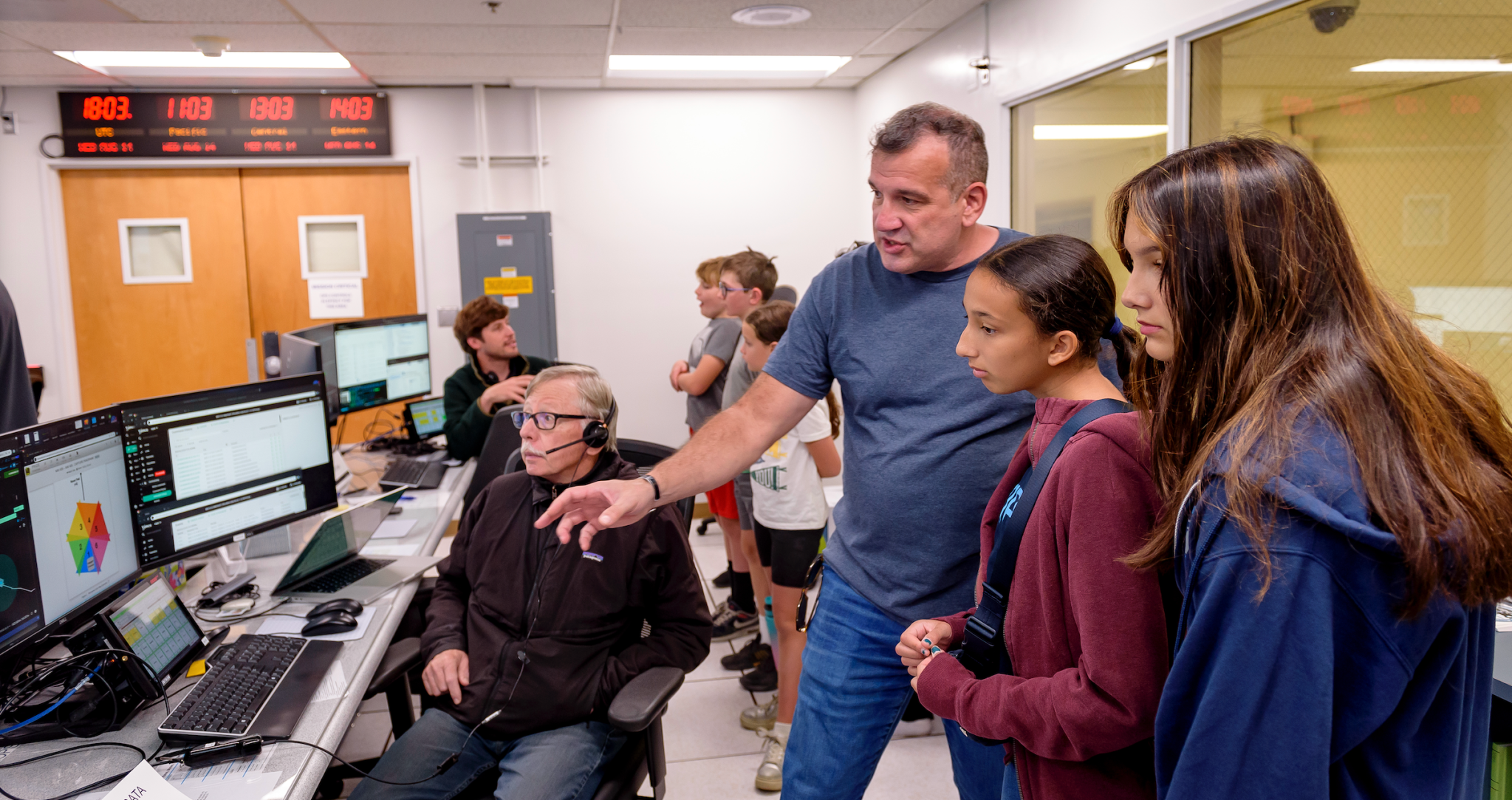 Chris Provencher explains the Volatiles Investigating Polar Exploration Rover (VIPER) mission to his family in the Multi-Mission Operations Center (MMOC), N240, for VIPER Friends and Family Day. Dennis Heher is seated at the control console at the left.