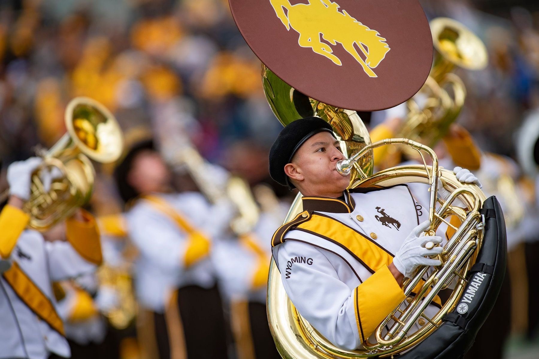 Aaron Vigil plays the sousaphone at University of Wyoming in Laramie. He wears marching band gear in white, gold, and maroon. Other band members are blurred, but visible in the background.