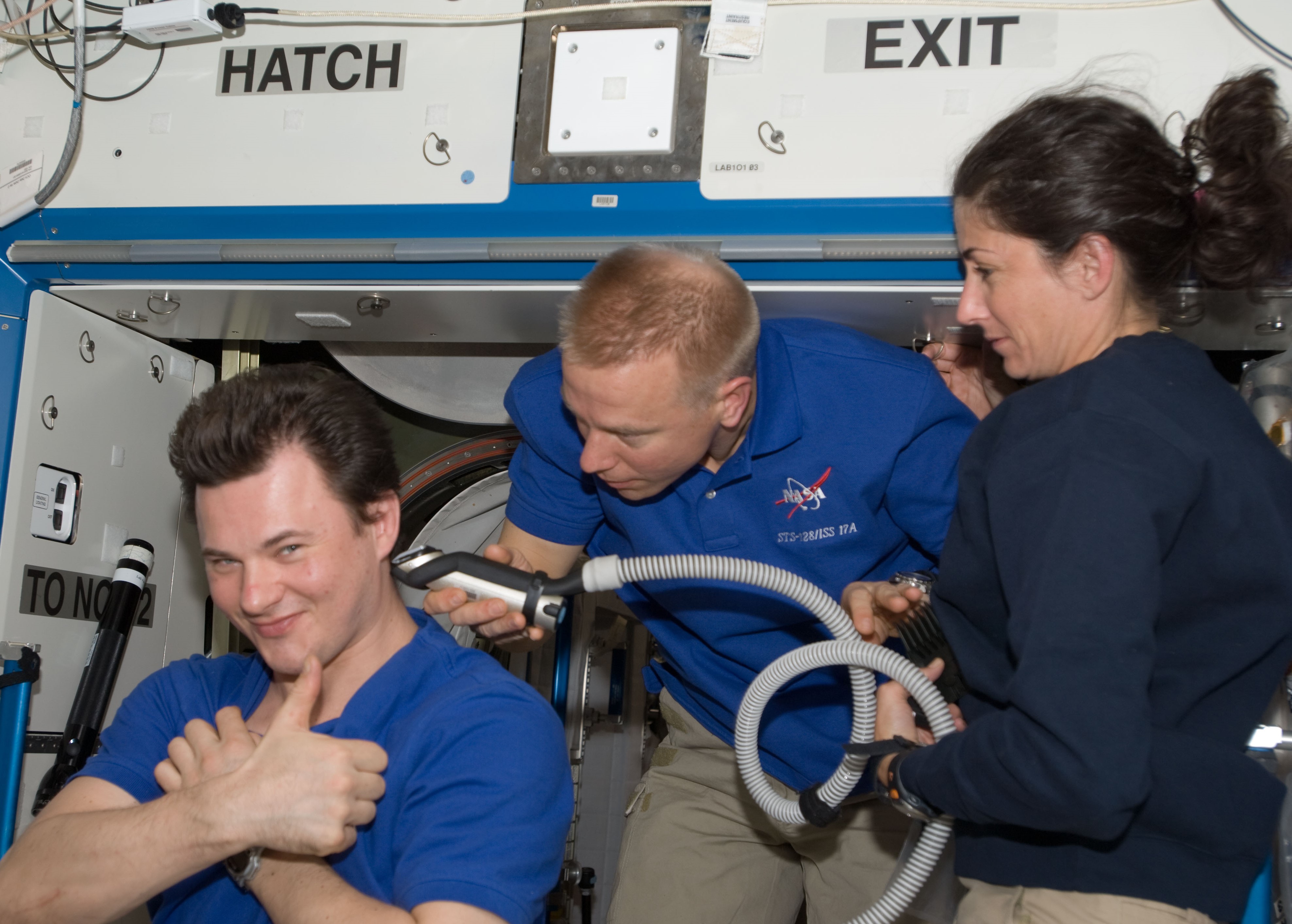 During handover operations, outgoing space station crew member Timothy L. Kopra, middle, shows incoming crew member Nicole P. Stott how to give a proper haircut in space