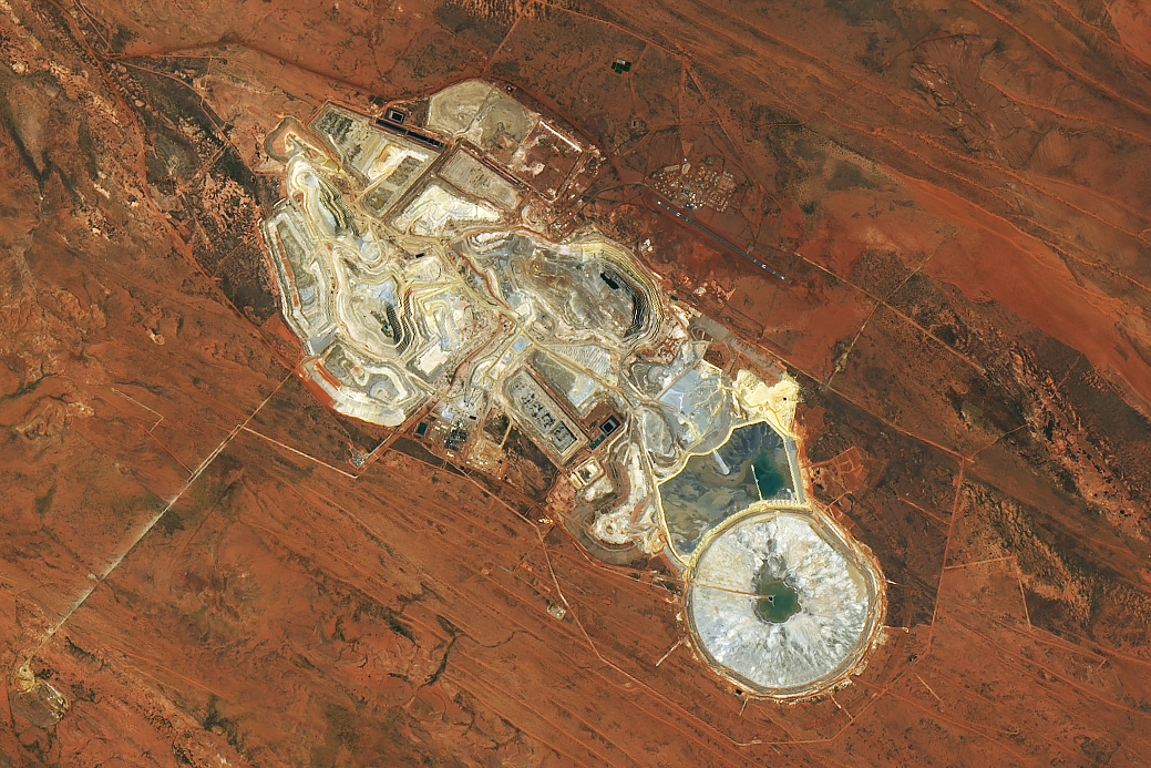 An overhead view of a mine in the dry outback of Australia. Most of the image is dominated by rusty red soil. Miners have excavated the soil in places, revealing an off-white color in these open pits. Some water is visible as well, in ponds.