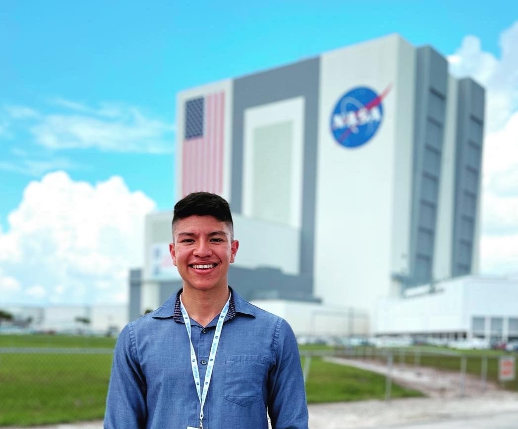 Aaron Vigil stands in front of building with American Flag and NASA meatball. He wears a blue button down shirt. 