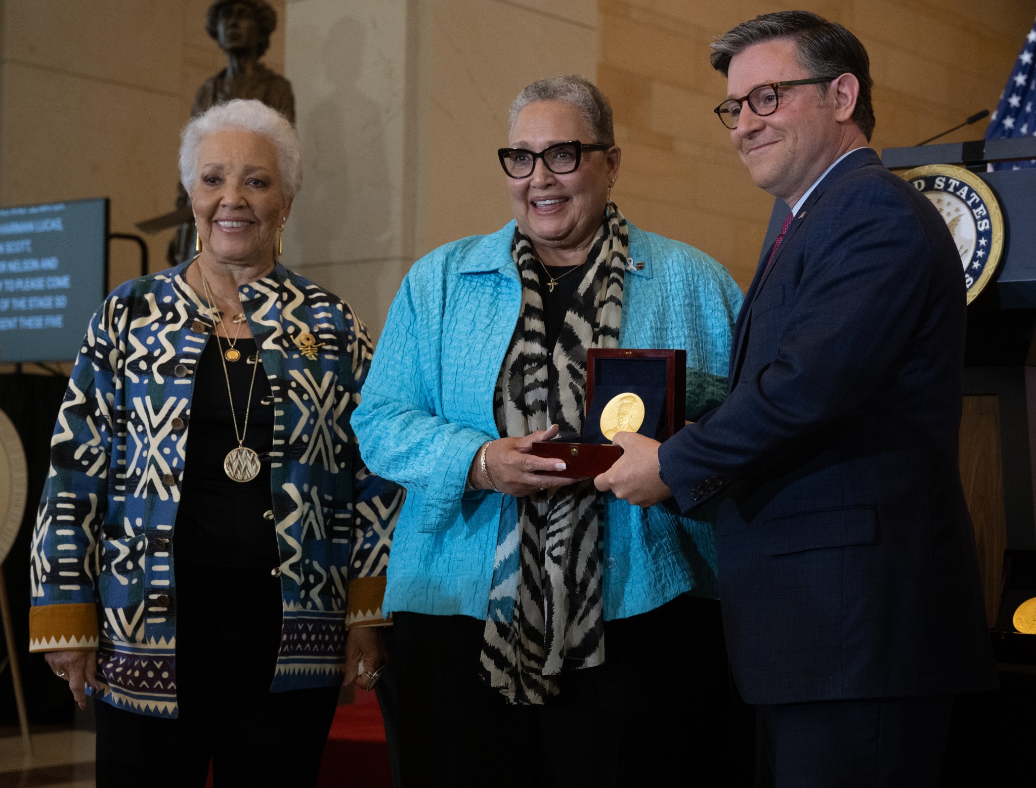 Two Black women and a white man face slightly left as they pose for a photo. The man and the woman in the middle hold a box containing a Congressional Gold Medal.