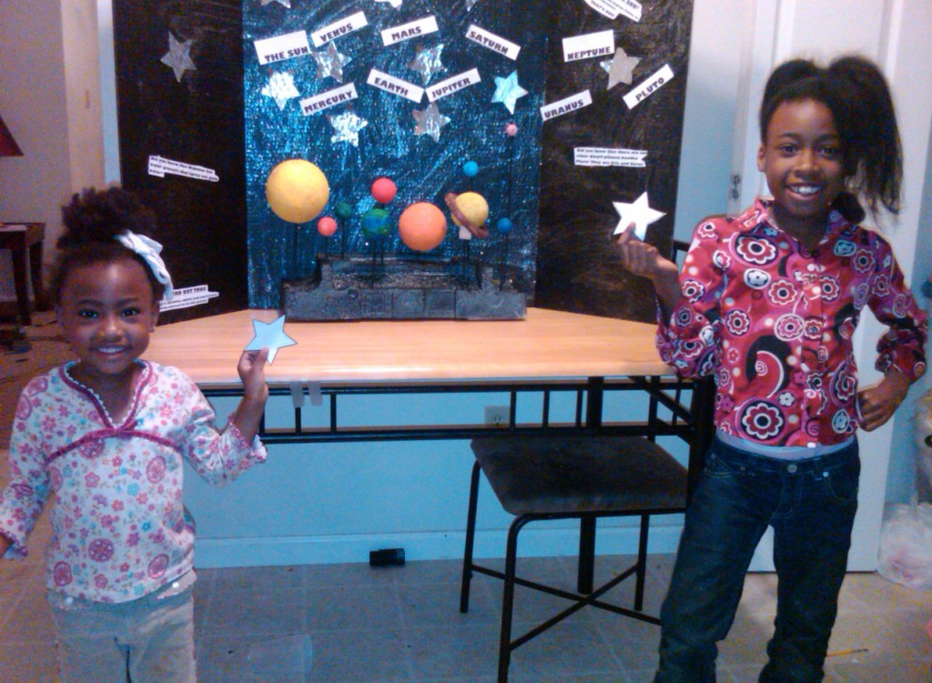 NASA intern Caitlyn McClanahan as a young student posing with her younger sister Erin in front of a science fair project, a recreation of the solar system. The two smile holding paper cutouts of stars. Credit: Caitlyn McClanahan