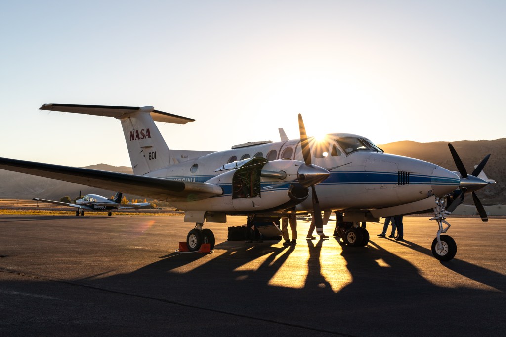 The outline of a small white plane with a blue stripe is mostly silhouetted in the gold light of a setting sun.