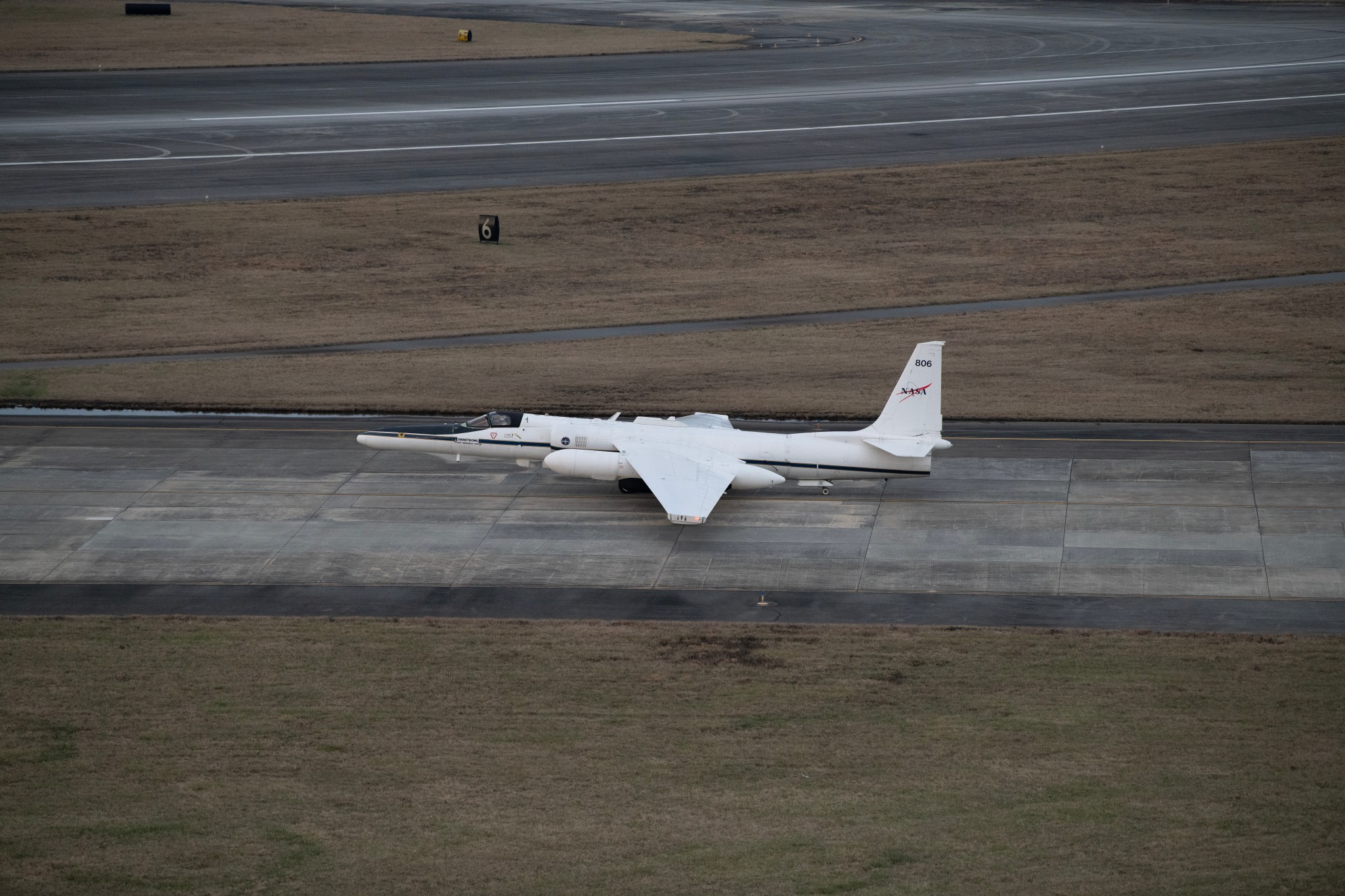 An aircraft is centered in the image facing the left. It is on a runway of dark gray concrete and the surroundings are primarily dry grass, with another runway at the top of the image.