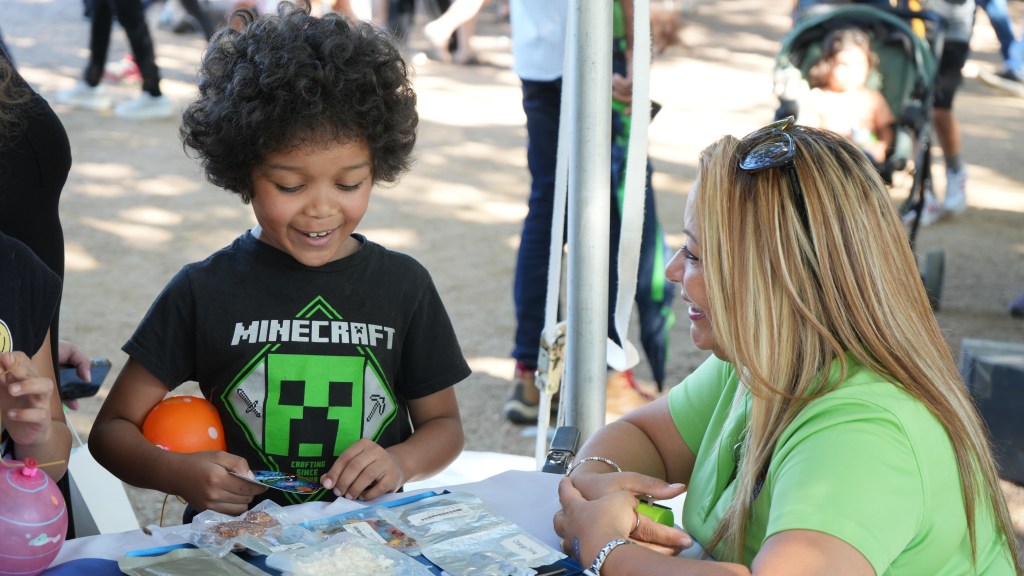 A smiling child wearing a Minecraft T-shirt explores a display of space-themed items, including packaged astronaut food, while a woman sits at the table next to him.