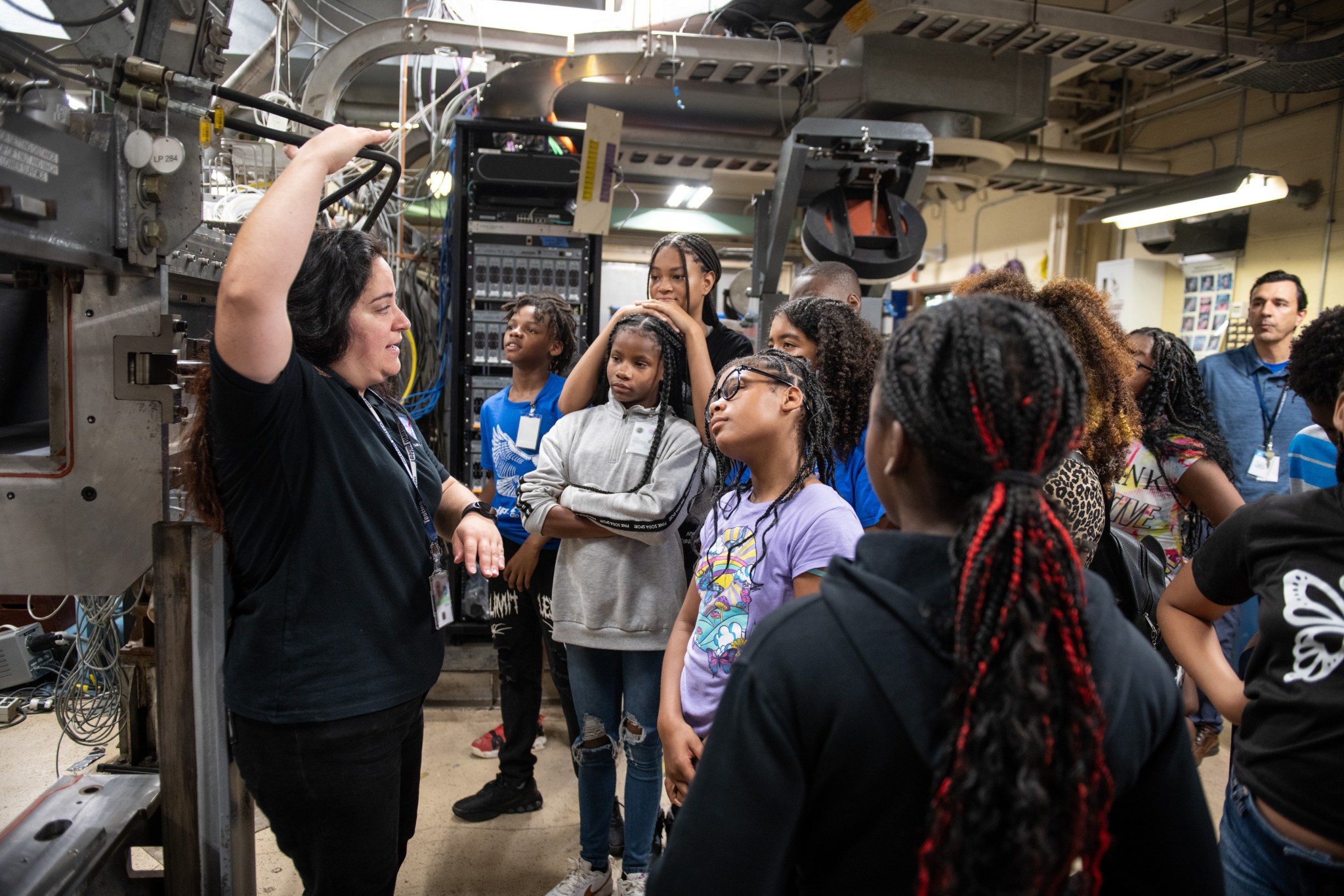 An engineer addresses a group of predominately female students in front of a test unit in a test facility filled with test hardware.