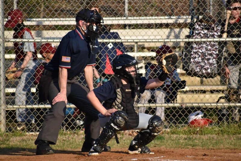 In front of a fence, Dr. Kenyon is dressed in an umpire uniform with a mask and stands behind a catcher. People sit in stands on the other side of the fence.