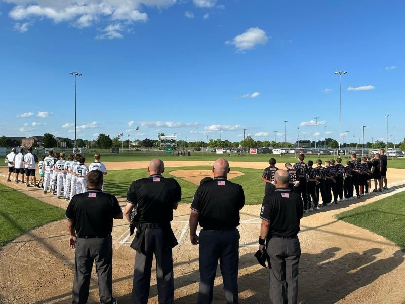 Four umpires, including Dr. Kenyon at the far right, stand with hand on heart for the national anthem. They stare down the middle of the field, flanked by two teams of players in front.