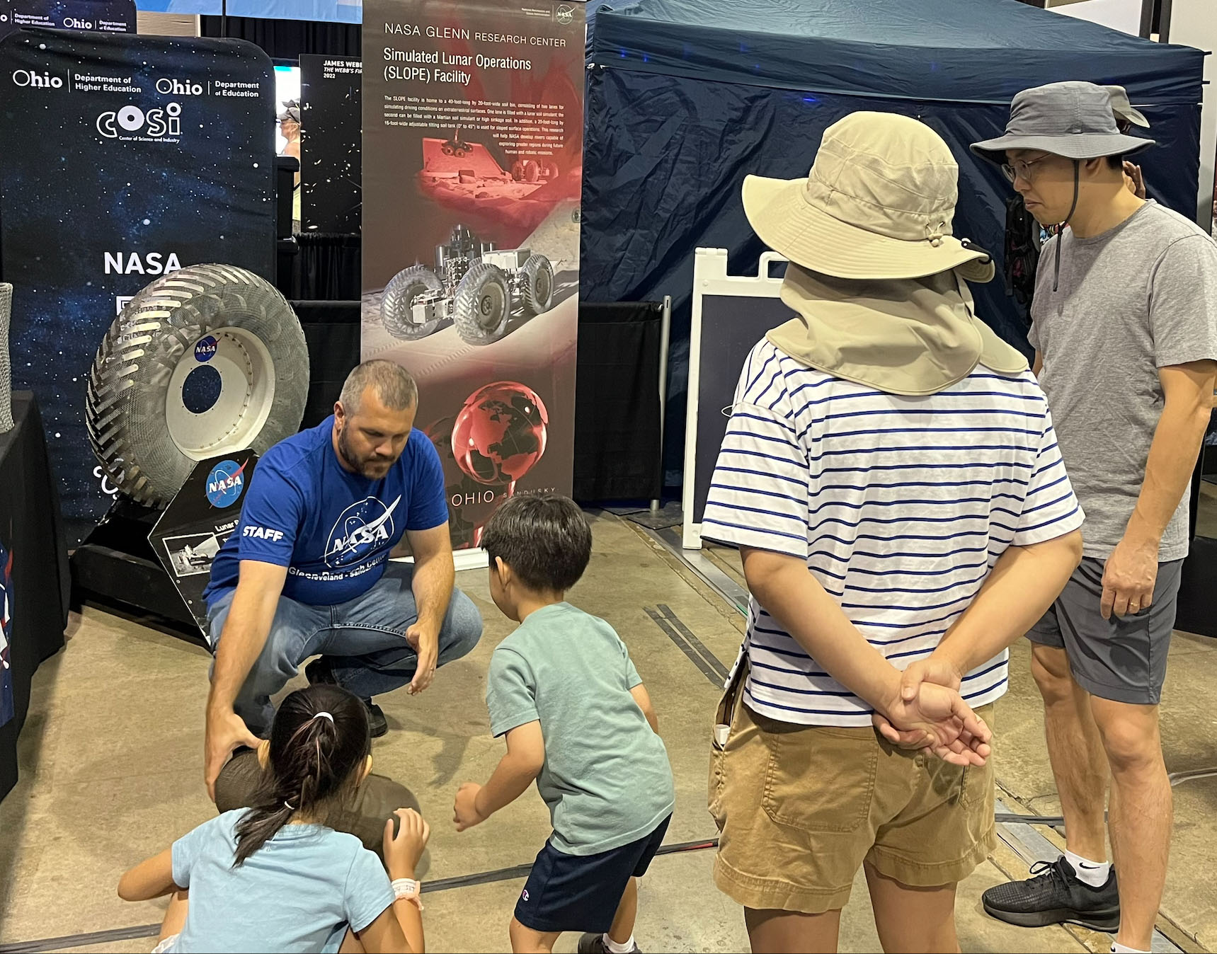 In front of colorful backdrops of tires, a NASA outreach specialist shows two children tires designed for Moon and Mars terrains. Parents stand to the right.