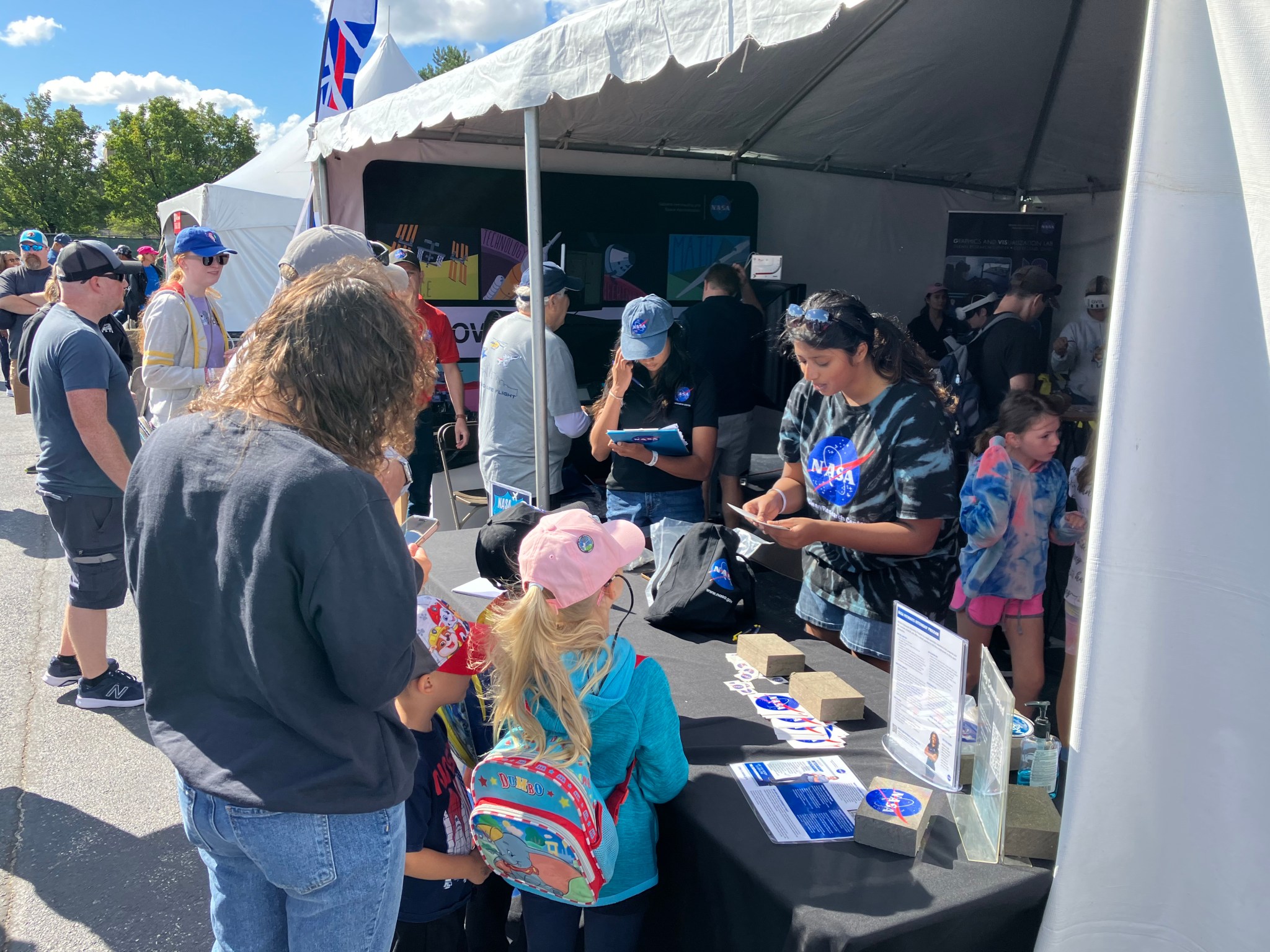 Two interns stand behind a table at the edge of a tent and interact with guests. Several other people wait to go inside the tent filled with interactive technology.