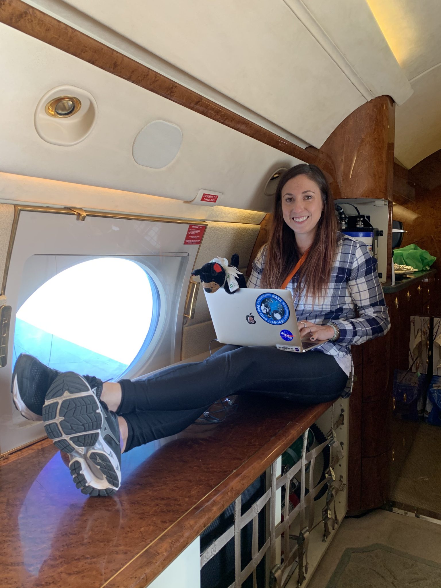 Linette Boisvert sits on a bench inside of an airplane with her laptop on her lap. She is wearing a blue and white plaid shirt, black pants, and tennis shoes.