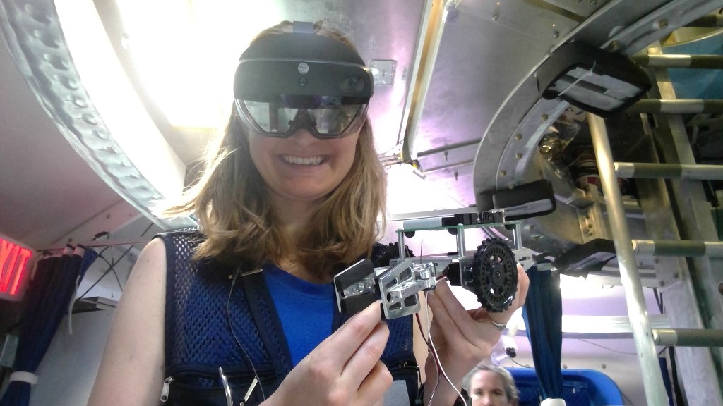 A person smiles while wearing an augmented reality headset and holding a small mechanical device inside HERA.