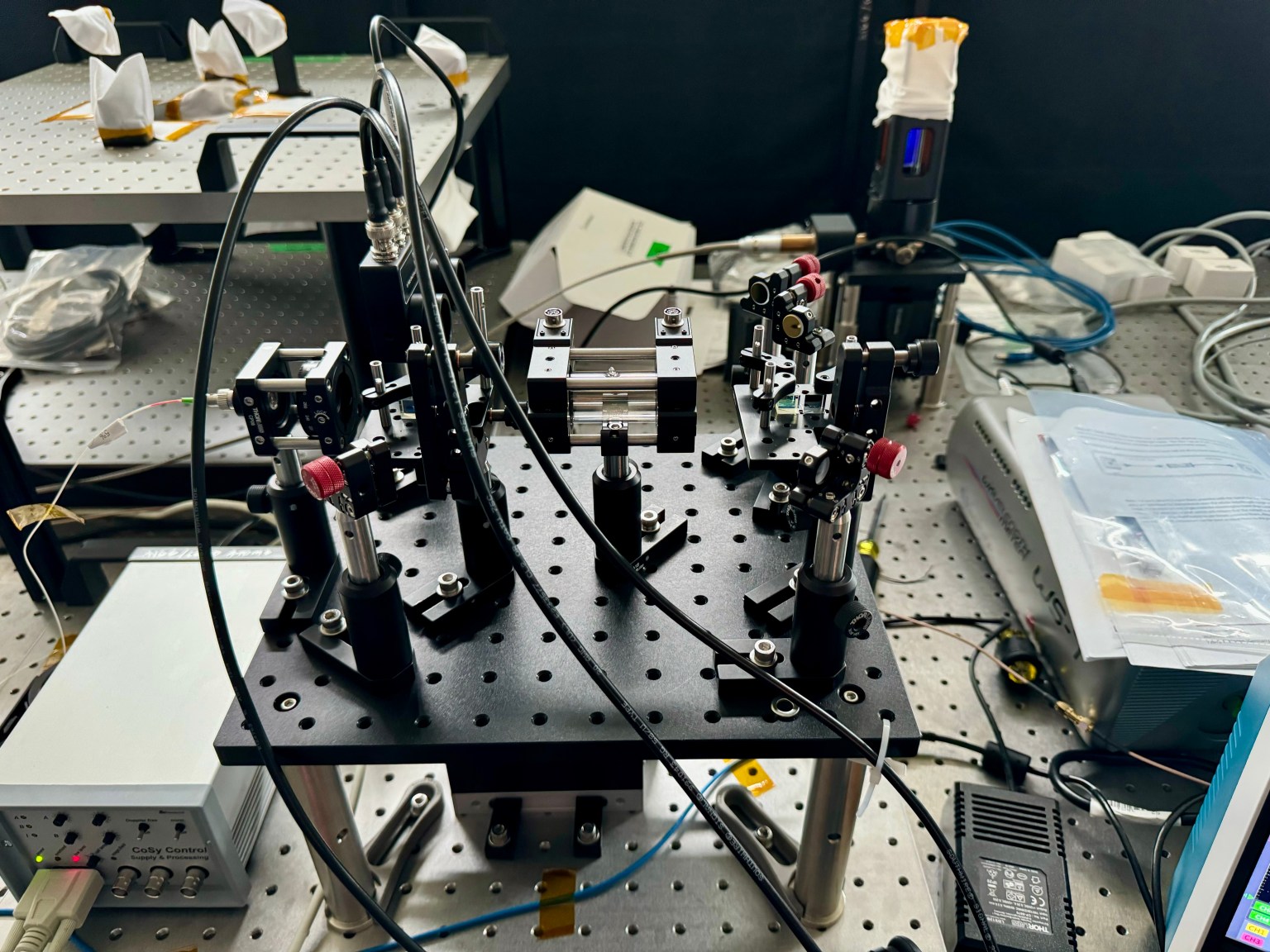 This image shows the Optical Atomic Strontium Ion Clock (OASIC), a small clear rectangular prism with wires and tubes surrounding it. The device is placed on a cluttered laboratory table, with other scientific instruments scattered about.