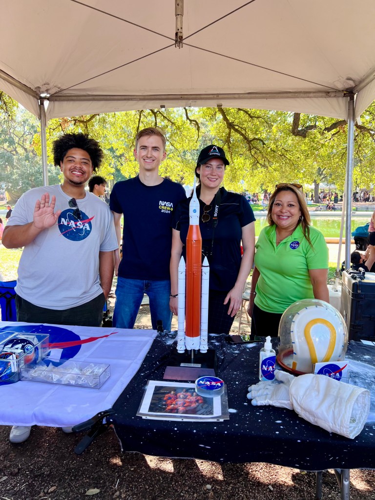  A group of four people stand behind a NASA booth table at an outdoor event. 