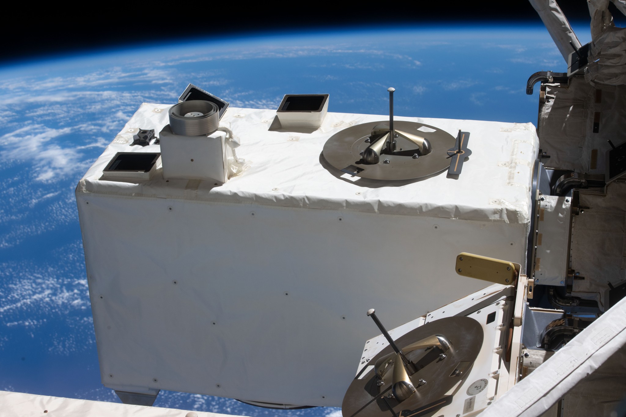 Exterior view of a white scientific instrument attached to the outside of the International Space Station, with Earth visible in the background.