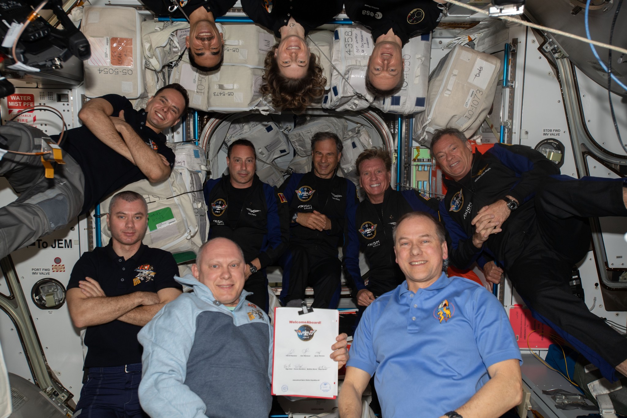 All 11 crew members are facing the camera and smiling. Artemyev is wearing a black polo shirt, Mateev a long-sleeved blue and white shirt and Korsakov a blue polo shirt. The rest of the crew members are wearing black or dark blue polo shirts. The three astronauts in the back row are upside down in relation to the others.