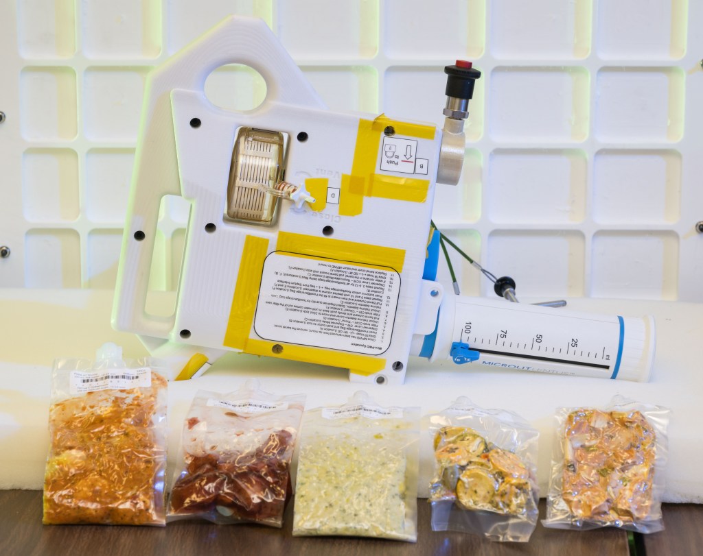 The image shows a compact, handheld water dispenser device placed on a table, with several vacuum-sealed packets of food arranged in front of it. The device is white with yellow tape and has various labels and instructions on its surface. It includes a large, graduated syringe-like component marked in milliliters, used for precise water measurement. The food packets in front of the device contain different types of rehydratable meals in various colors and textures, indicating the preparation of food for consumption in a space environment. The background features a white grid pattern, enhancing the focus on the device and food packets.