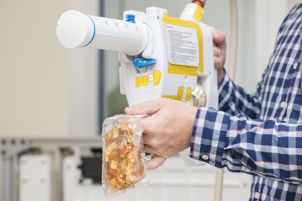 The image shows a close-up of an engineer's hands as they operate a Mini Potable Water Dispenser. The engineer is holding the white, handheld device, which features a large, graduated syringe-like component and is marked with various yellow labels and instructions. The engineer is rehydrating a vacuum-sealed food pouch, which he is holding in one hand while the other hand operates the dispenser. The food inside the pouch appears to be a mix of ingredients. The engineer is wearing a blue checkered shirt.