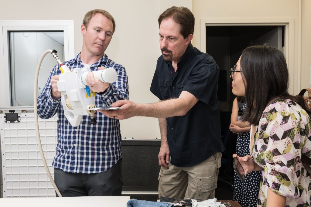 The image shows a group of three people engaged in a demonstration of the Mini Potable Water Dispenser. The person on the left, wearing a checkered shirt, is holding the device and showing its operation to the others. The person in the center is closely examining a vacuum-sealed food pouch being held by the person with the dispenser, while pointing or assisting in the demonstration. The person on the right, wearing glasses and a floral-patterned top, is attentively observing the process, holding a smartphone, possibly ready to capture the demonstration.