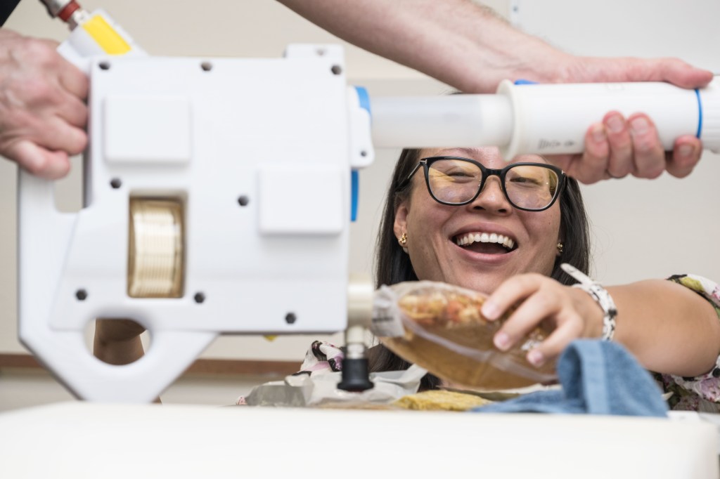 A NASA food scientist smiles as she rehydrates a food pouch using the Mini Potable Water Dispenser during a demonstration at Johnson Space Center. The scientist holds the food pouch steady while another person manually operates the handheld device, which is designed to dispense water for preparing meals and maintaining hygiene on the Gateway lunar space station.