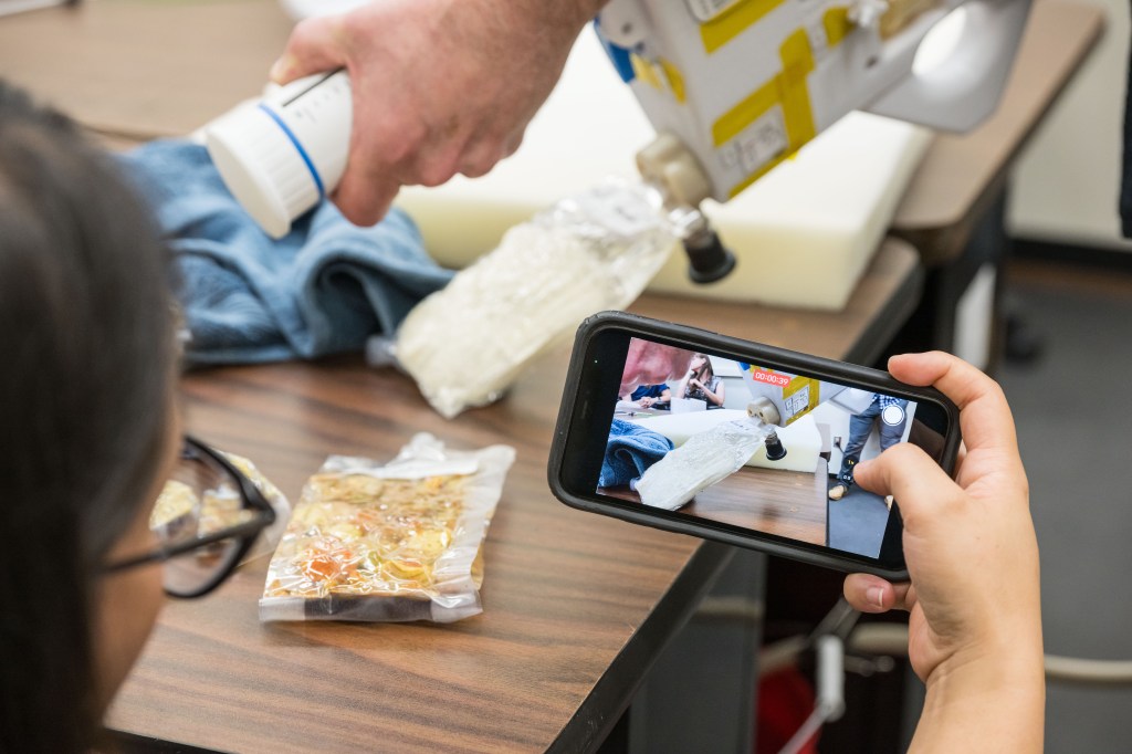 The image shows a close-up of a person filming a demonstration with a smartphone. The scene being recorded involves the use of a Mini Potable Water Dispenser to rehydrate a vacuum-sealed food pouch. The person's hand is holding the device while directing water into the pouch, which is positioned on a table. The smartphone's screen is clearly visible in the foreground, displaying a live video of the demonstration in progress