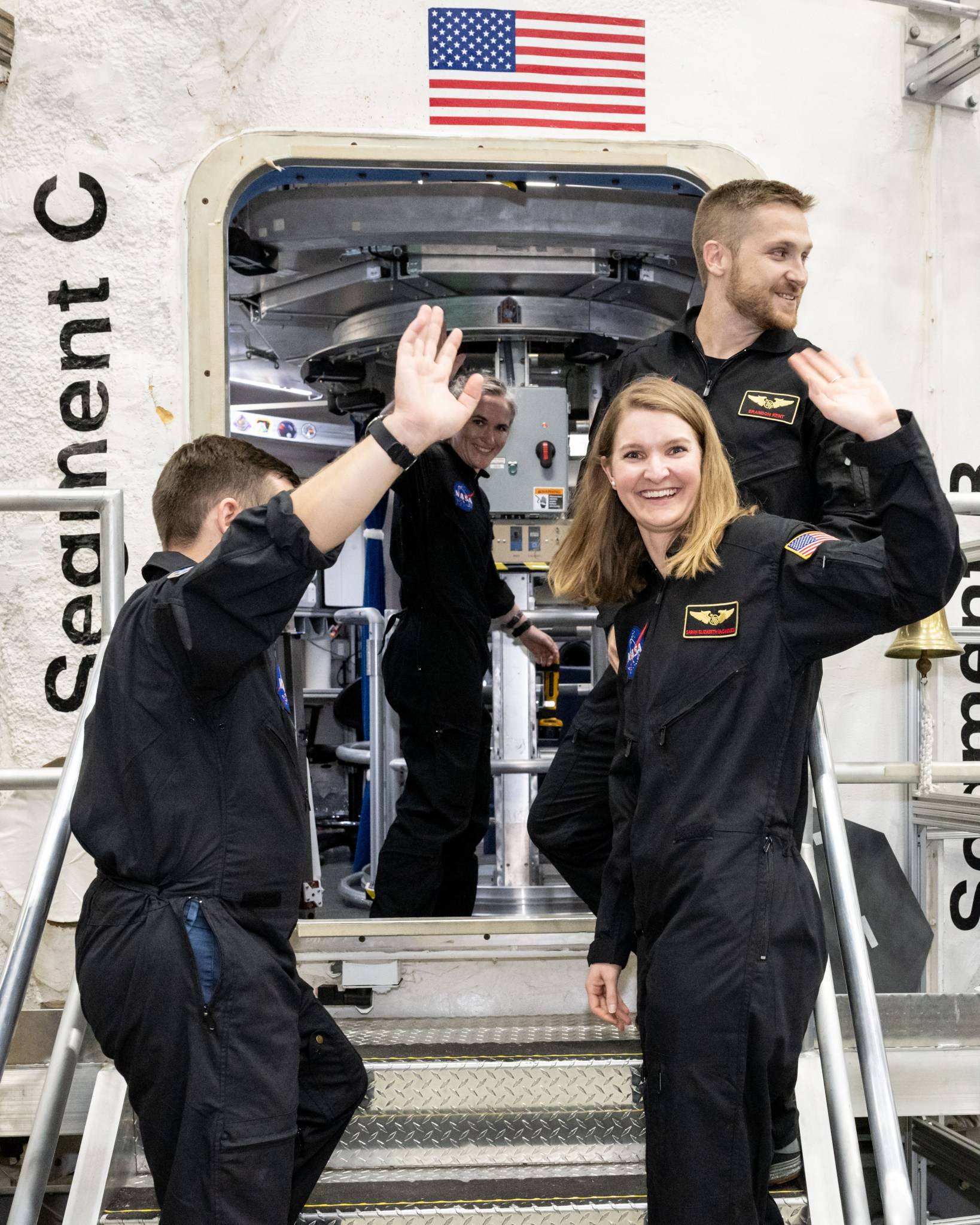 Four individuals in black NASA jumpsuits are smiling and waving as they stand on steps leading into the HERA habitat.