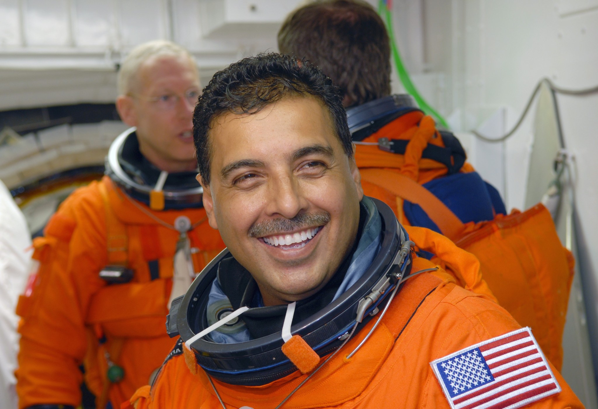 Astronaut José Hernández, a Hispanic man, smiles broadly at the camera while waiting to board space shuttle Discovery. He wears an orange escape suit that has an American flag patch on the left shoulder. Behind him are mission specialists Patrick Forrester (left) and Christer Fuglesang (back to camera).