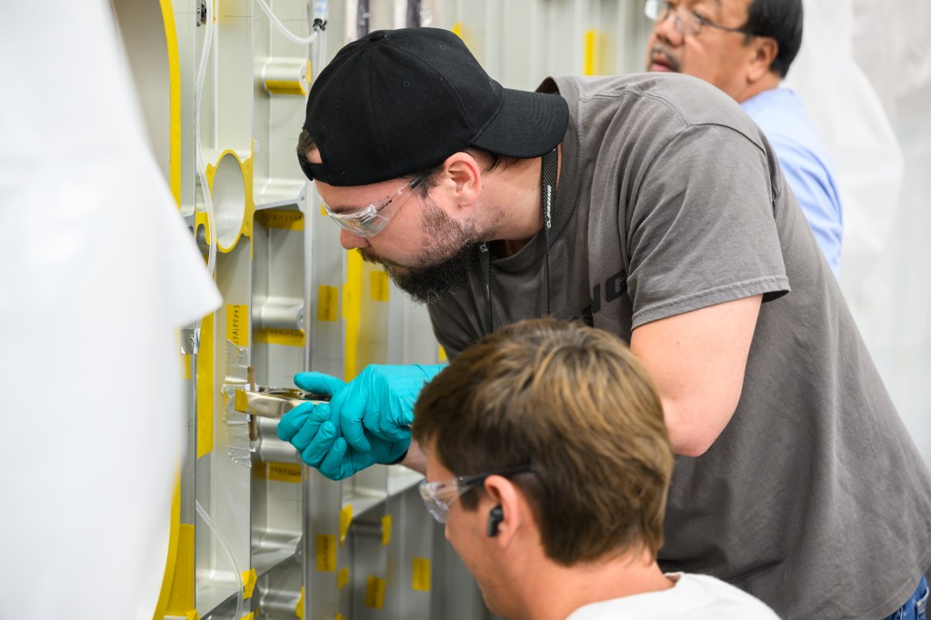 : NASA Michoud Assembly facility technicians Cameron Shiro (foreground), Michael Roberts, and Tien Nguyen (background) install the strain gauge on the forward adapter barrel structural test article for the exploration upper stage of the SLS rocket.