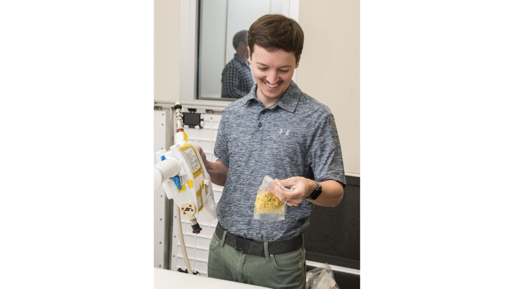 The image shows a person smiling as they try the Mini Potable Water Dispenser for the first time. The individual, dressed in a gray polo shirt and green pants, is holding the white, handheld dispenser in one hand while examining a vacuum-sealed food pouch in the other.