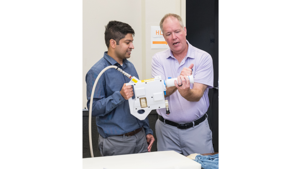 The image shows two individuals standing side by side as they demonstrate the Mini Potable Water Dispenser. The person on the left, dressed in a blue shirt, is holding the device, while the person on the right, wearing a light purple shirt, is explaining how to use it. The person on the right is gesturing with his hands, explaining the device's functionality or the process of rehydrating food. The device is connected to a hose and features a large, graduated syringe-like component.