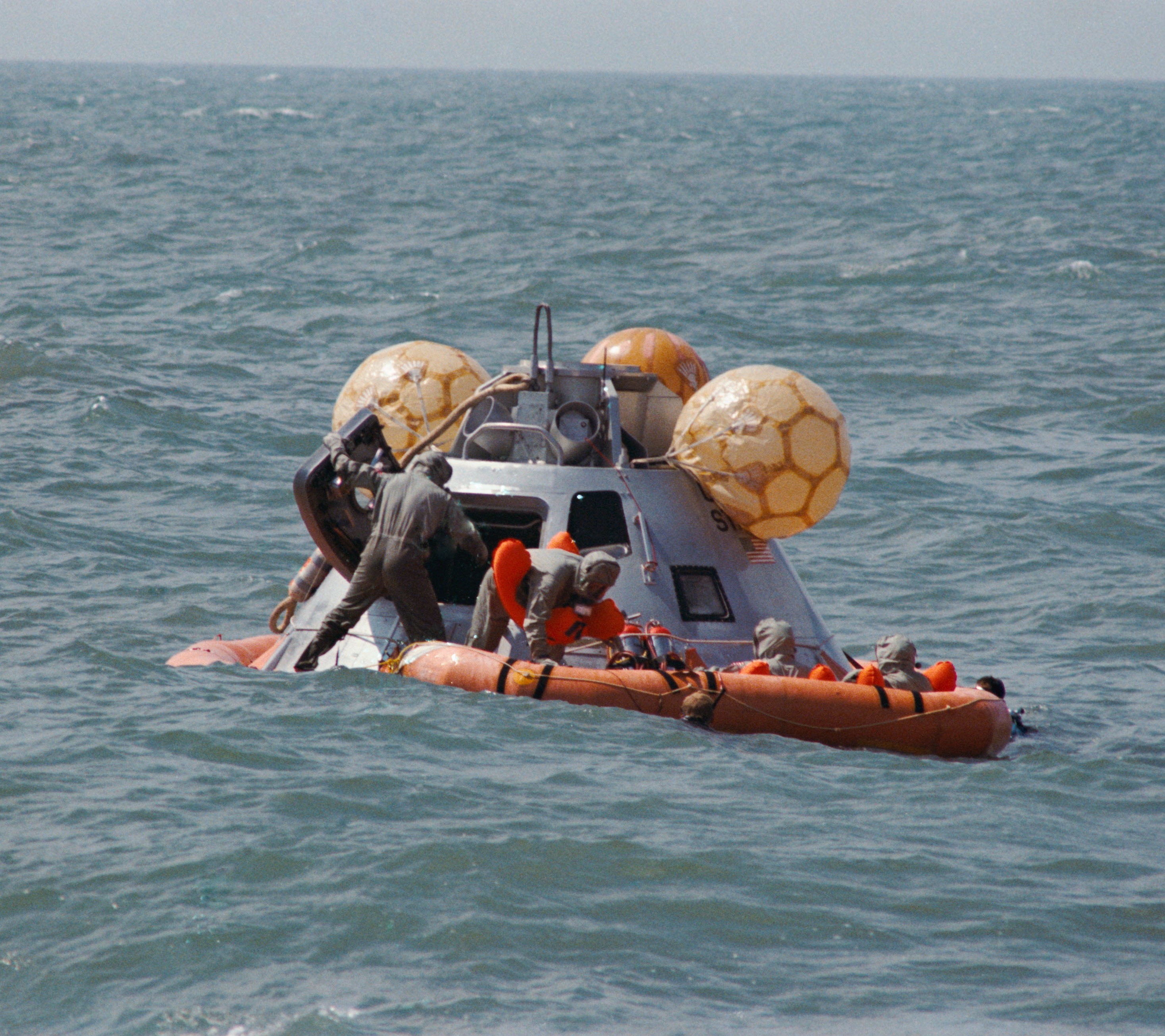 Wearing Biological Isolation Garments and assisted by a decontamination officer, standing in the open hatch, Apollo 12 astronauts await retrieval in the life raft