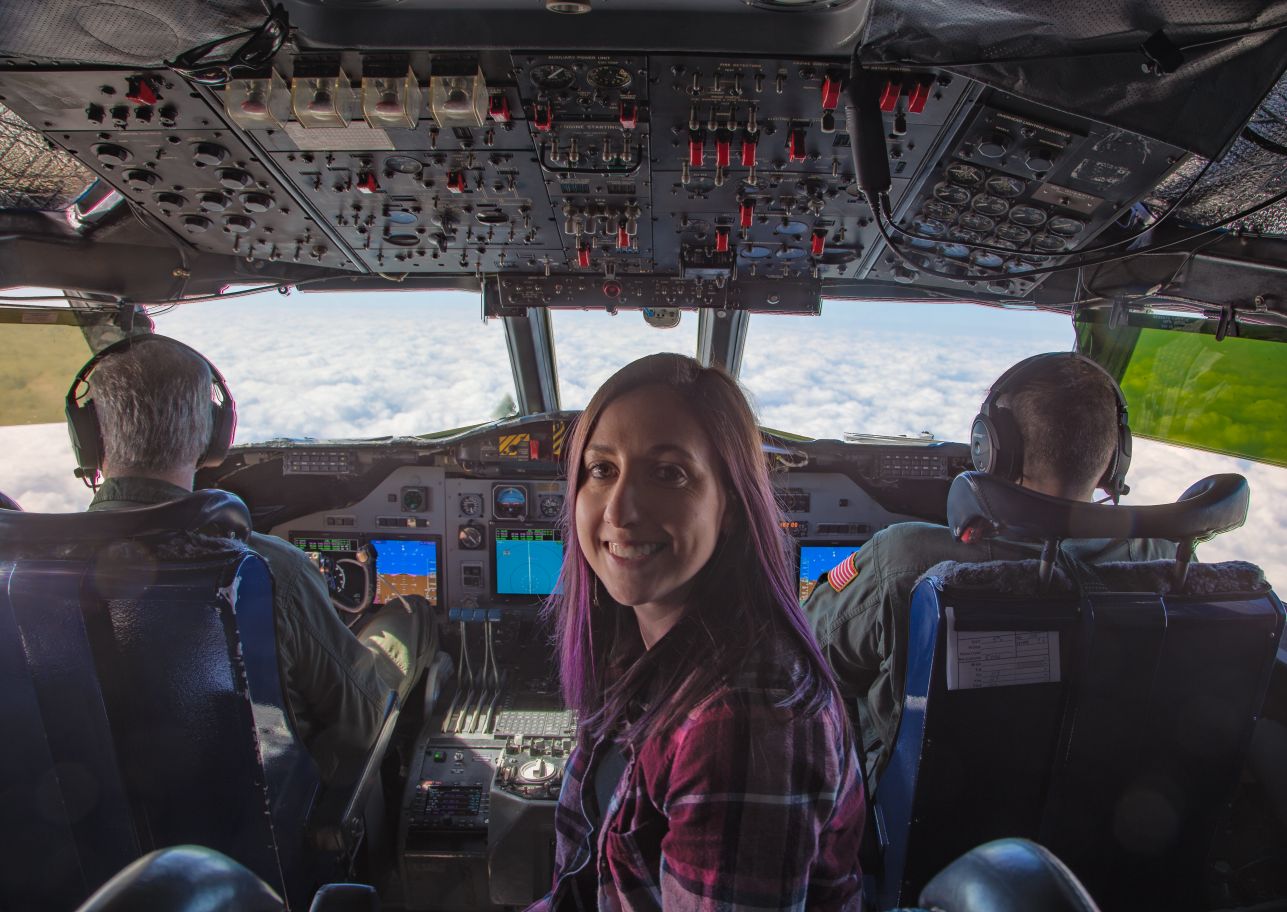 Linette Boisvert turns to smile at the camera as she sits in the center of a plane behind the pilots. Both pilots are visible along with the airplane controls. Through the front window of the plane, clouds are visible. 