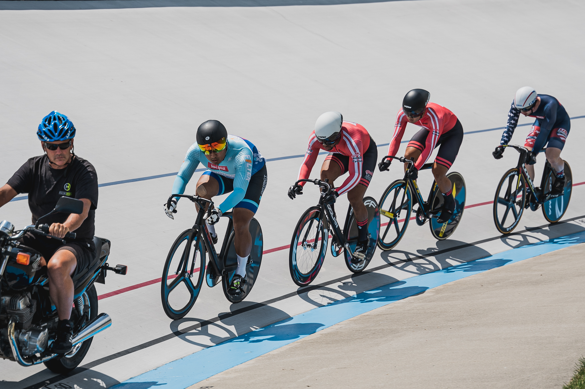 Four men on racing bikes during a keirin race on a track. They are in a single file line behind a man on a motorized bike wearing a blue helmet.