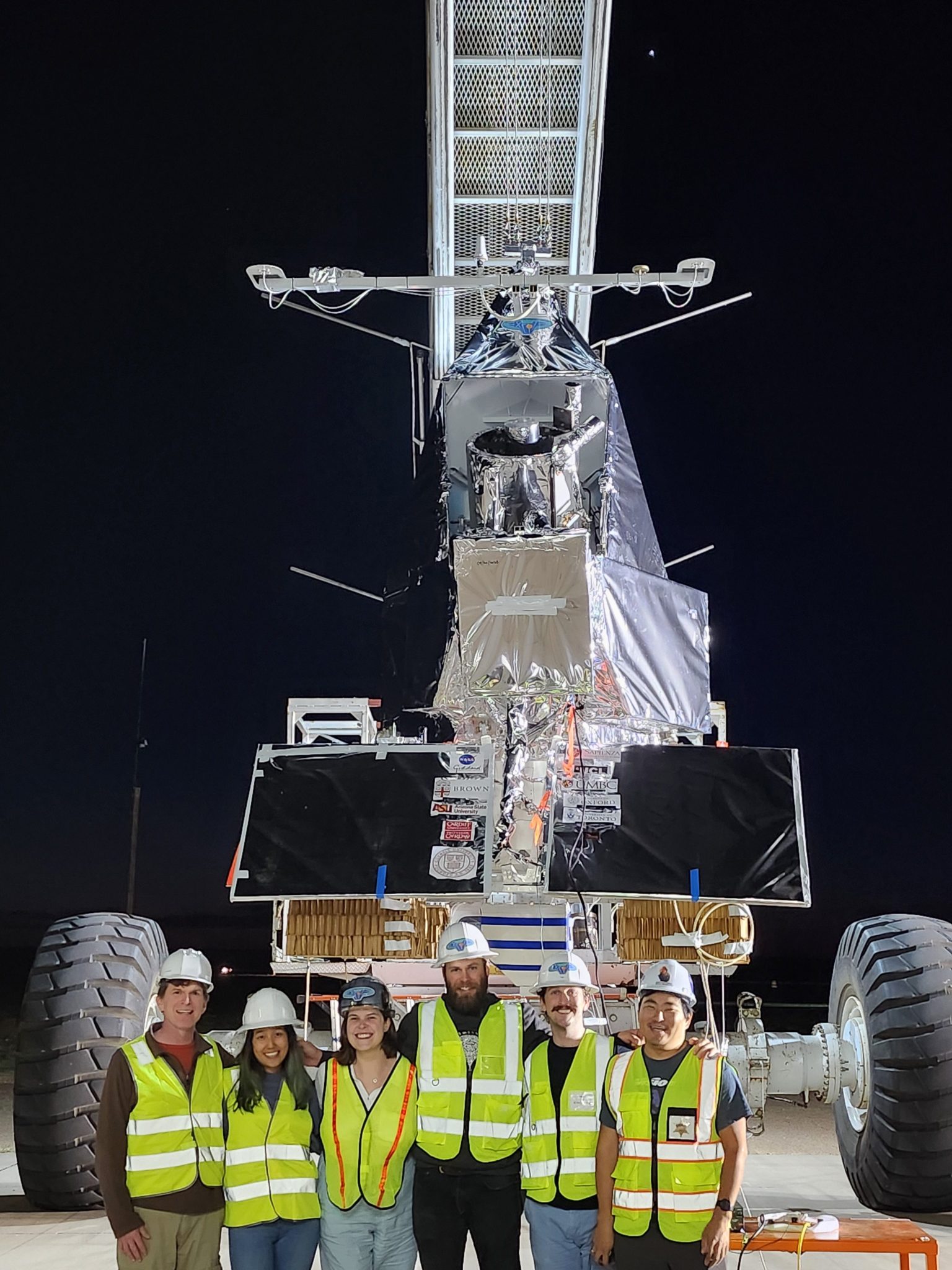 Six people wearing hard hats and yellow safety vests stand in front of a large spacecraft on a crane with large wheels on either side. 