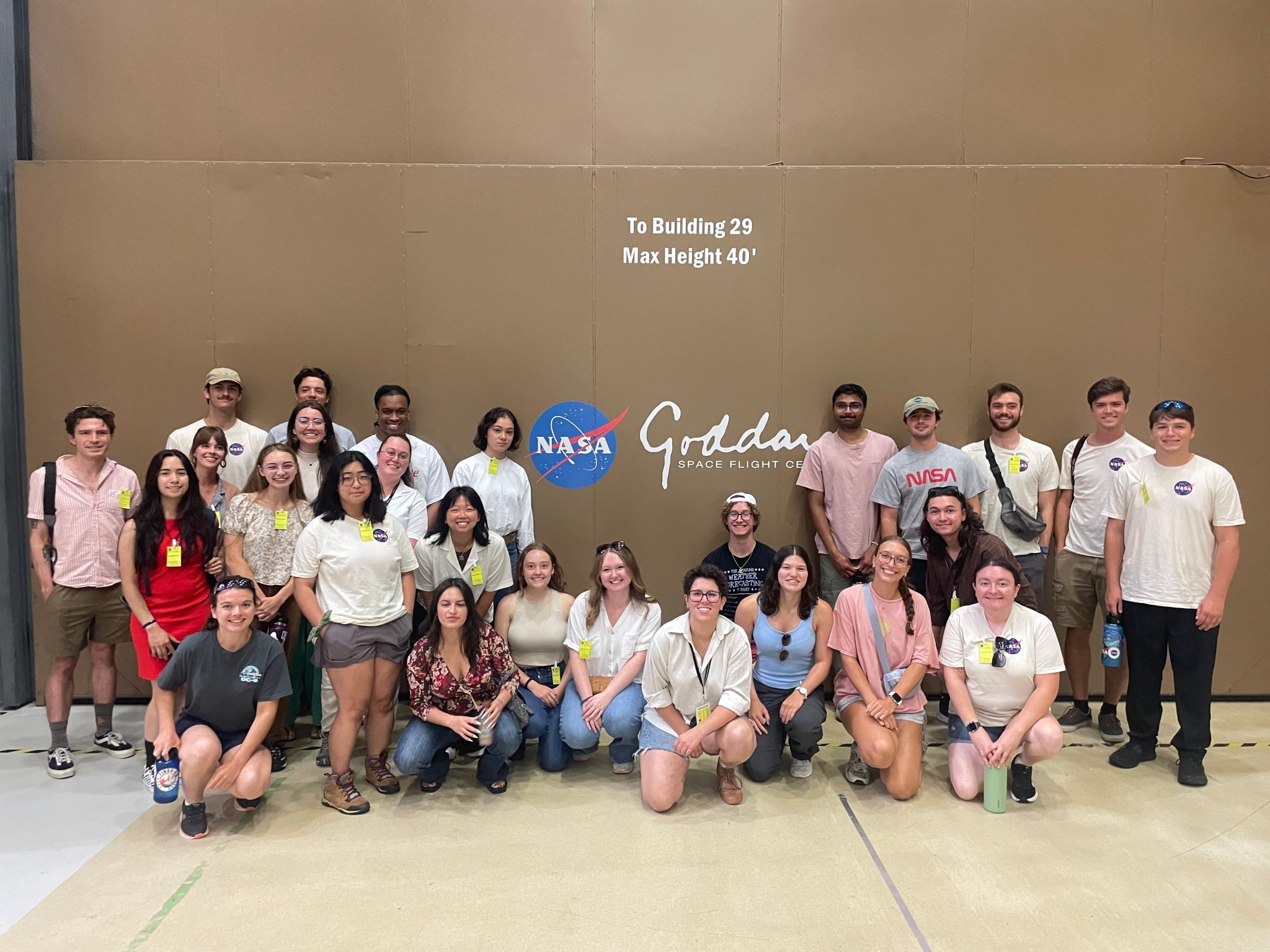 A group of about 25 interns stands in front of a brown wall that has the NASA logo and the word Goddard in white text.