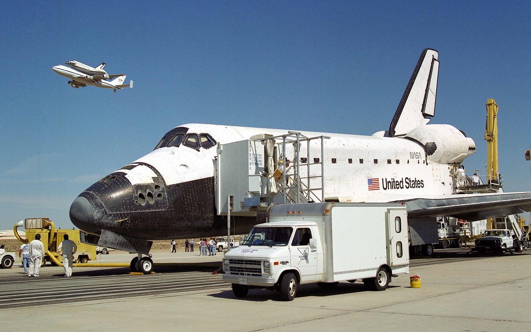 As workers process Endeavour on the runway, Columbia atop a Shuttle Carrier Aircraft (SCA) flies overhead on its way to the Palmdale facility for refurbishment